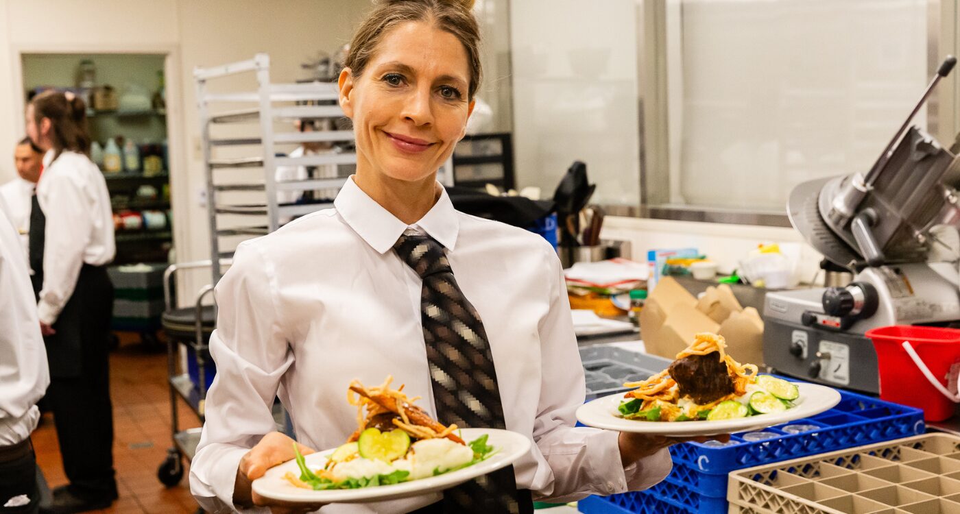 Trainee dressed in white shirt and tie carries two plates for diners at Fresh Starts Chef Events. She is smiling at the camera.