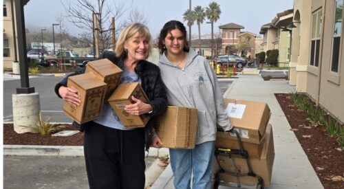 Older woman with blond hair holds three boxes. Next to her, a young woman with dark hair and sweatshirt pulls a handtruck full of boxes.