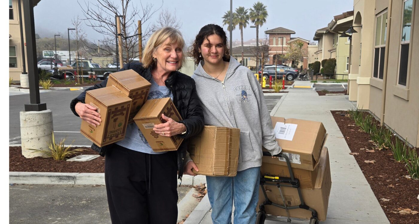 Older woman with blond hair holds three boxes. Next to her, a young woman with dark hair and sweatshirt pulls a handtruck full of boxes.