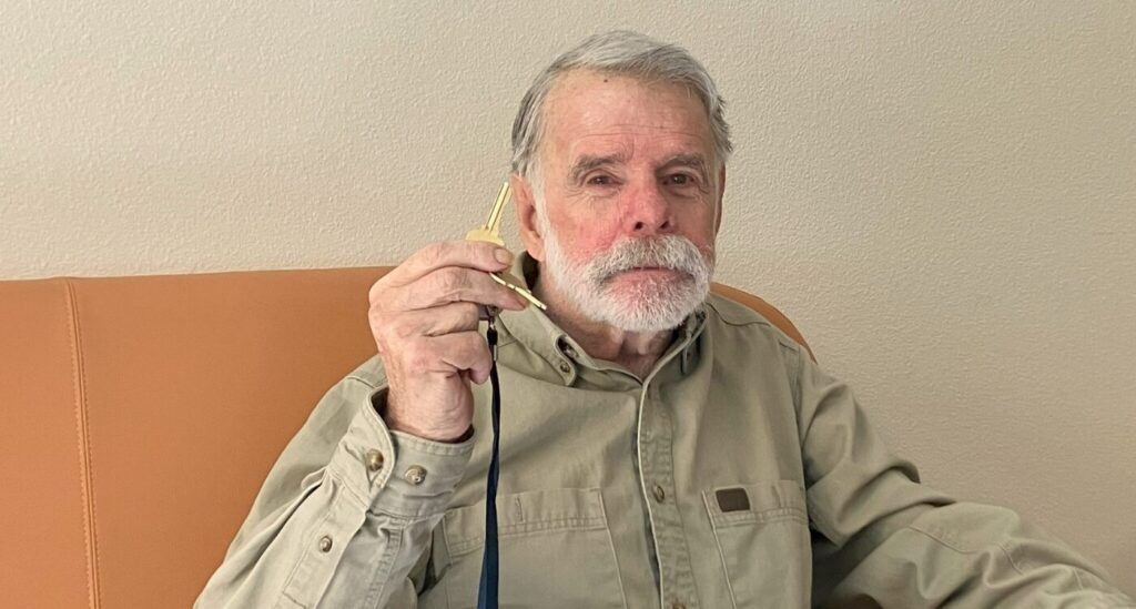 Veteran John M. sits on sofa in his new apartment holding up his keys with a smile.