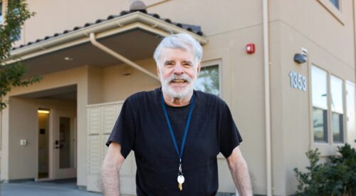 Man with white hair and beard smiles at the camera. He is wearing a lanyard of keys around his neck.