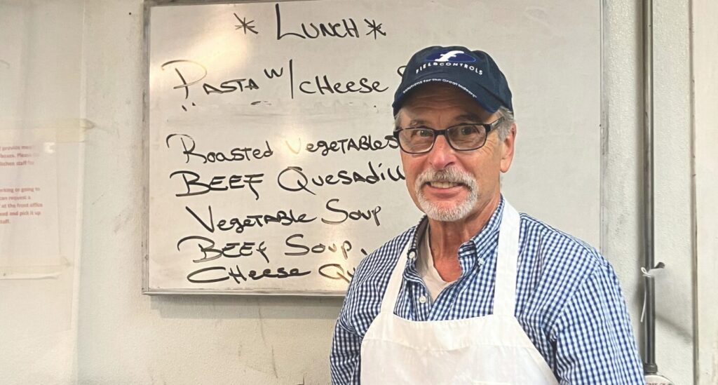 Man with beard wearing glasses and baseball cap stands in cafeteria-style kitchen. He smiles wearing button-down shirt and white apron.