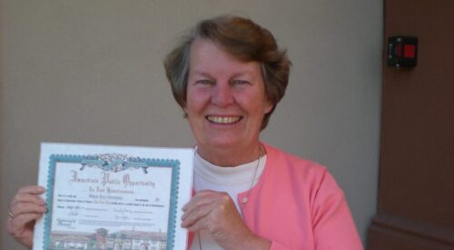 Short-haired woman in pink sweater holds a certificate and smiles at the camera.