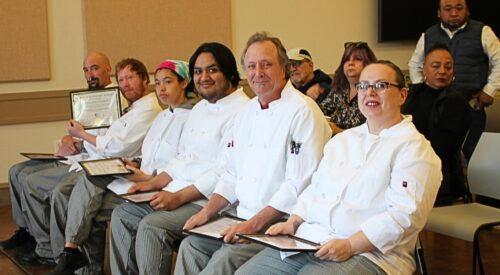 Six culinary students in white coats sit in the front row at the graduation event.