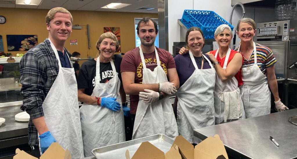 Four women and two young men wearing white aprons stand in commercial kitchen,