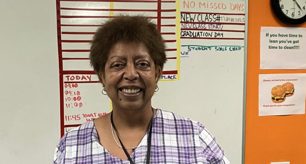 Black woman with short hair stands in a classroom and smiles at the camera