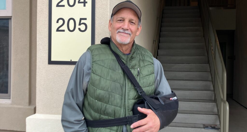 Man in baseball cap and green vest stands by apartment building.