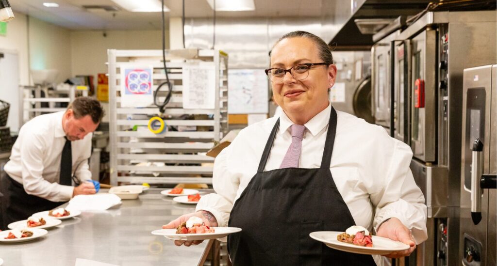 Woman with glasses carries plates. She is wearing a white shirt, tie and black apron.