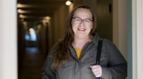 Woman with long brown hair and glasses smiles as she stands at doorway.