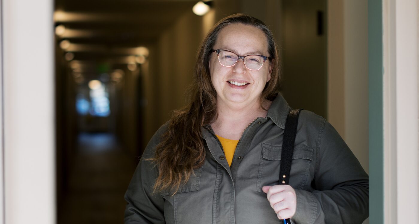 Woman with long brown hair and glasses smiles as she stands at doorway.