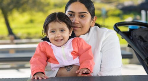 Mother with dark hair and white sweater hugs toddler daughter at a picnic table