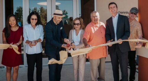 Seven people with scissors cut a gold ribbon in front of glass doors.