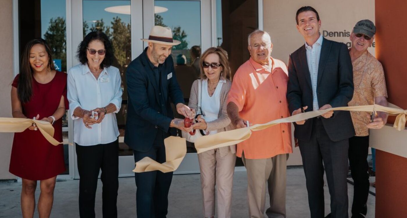 Seven people with scissors cut a gold ribbon in front of glass doors.