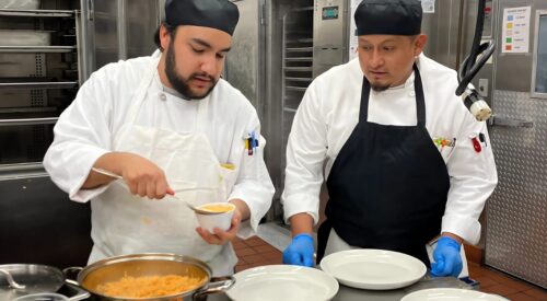 Two men in chef coats and aprons work in kitchen