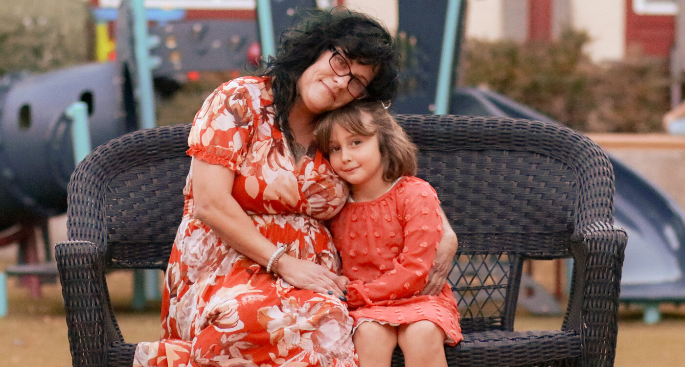 A mother and daughter sit on a wicker bench in front of a play structure.