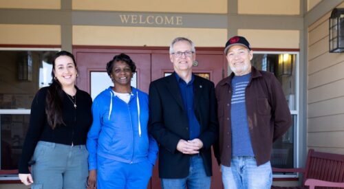 Two men and two women stand at the door of a building. A sign over the door says Welcome.