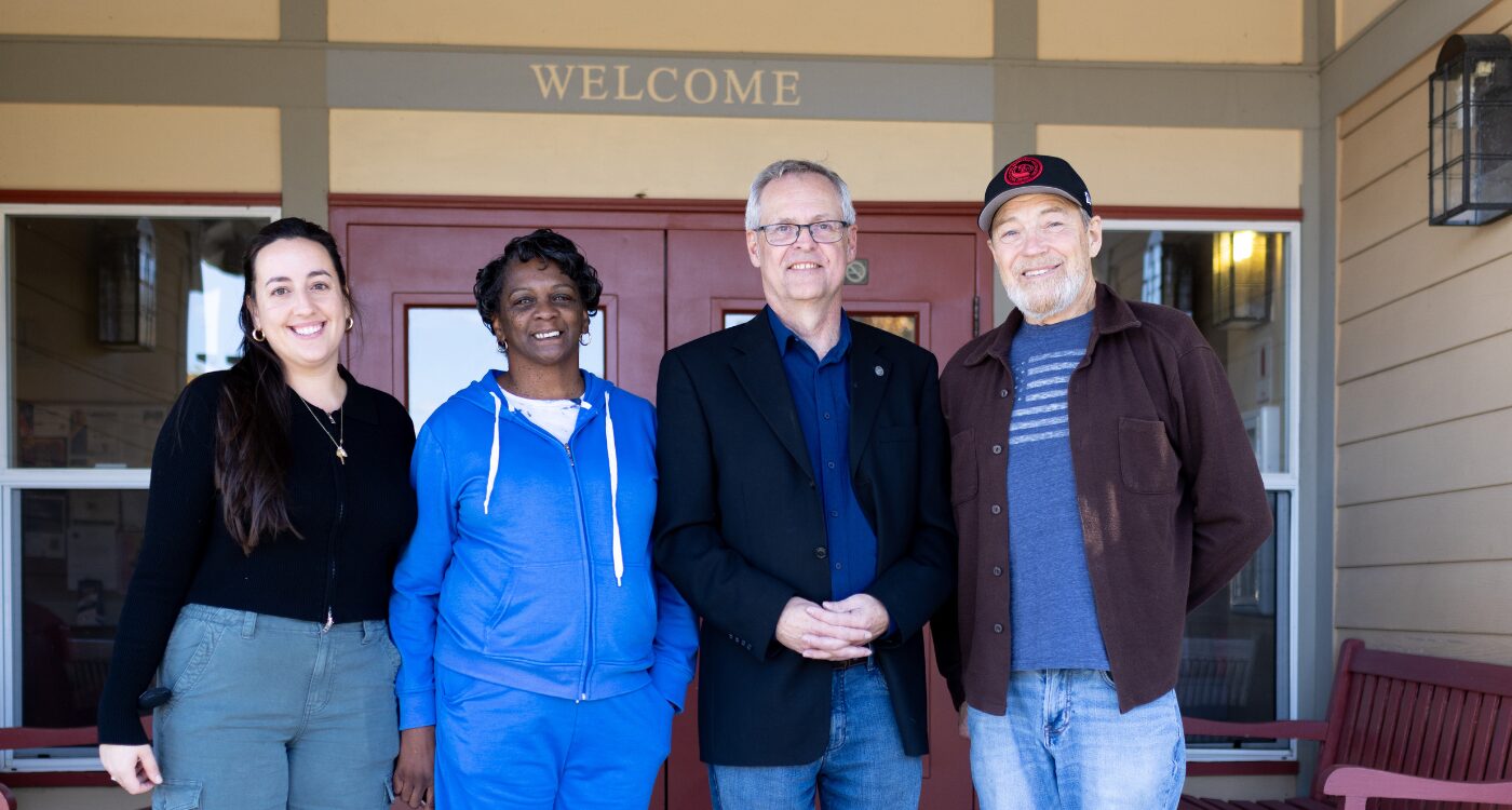 Two men and two women stand at the door of a building. A sign over the door says Welcome.