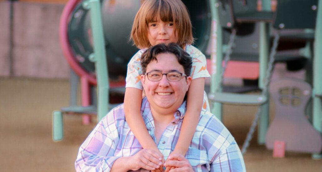 Woman with short dark hair and glasses kneels and holds hands of girl behind her