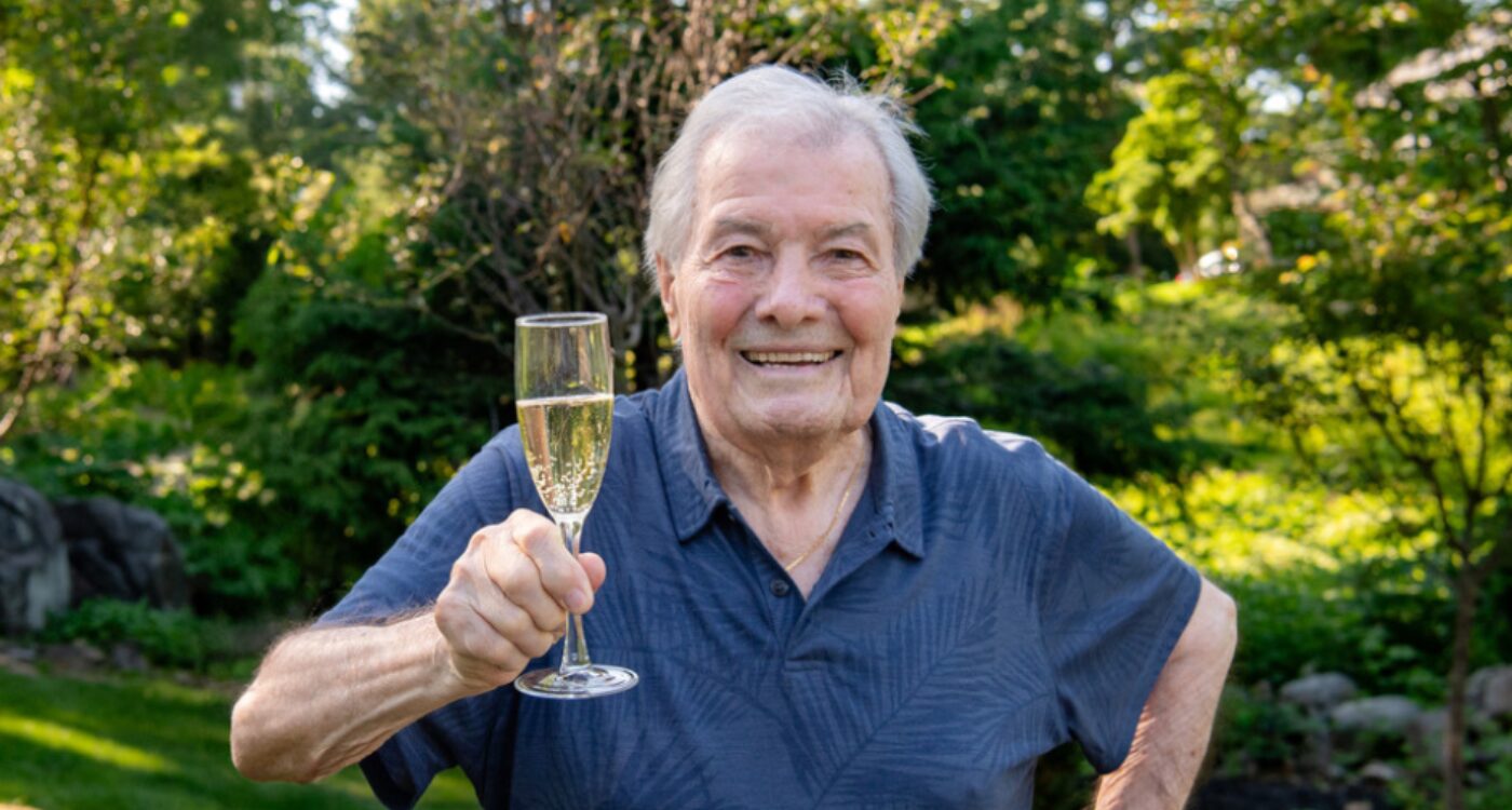 White-haired man in blue shirt holds glass of champagne