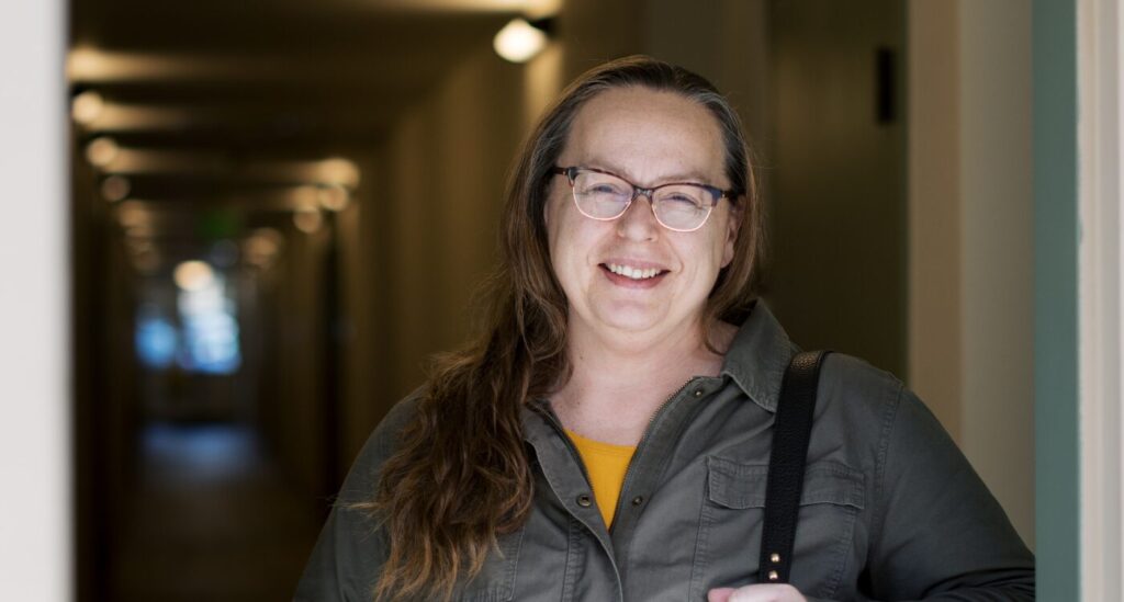 Woman with long hair and glasses smiles while standing in a doorway.
