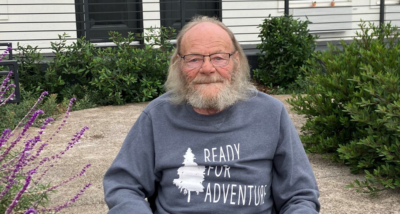 Older man with glasses and blue sweatshirt sits in garden