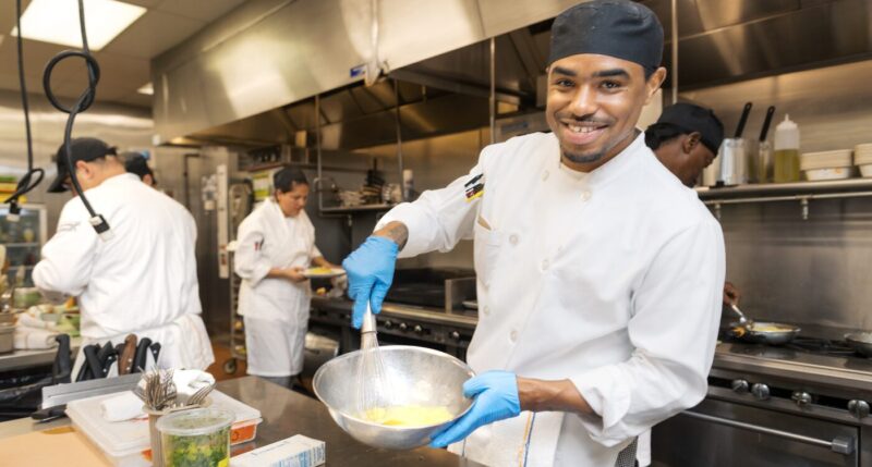 Man in white chef coat smiles while whisking bowl of eggs