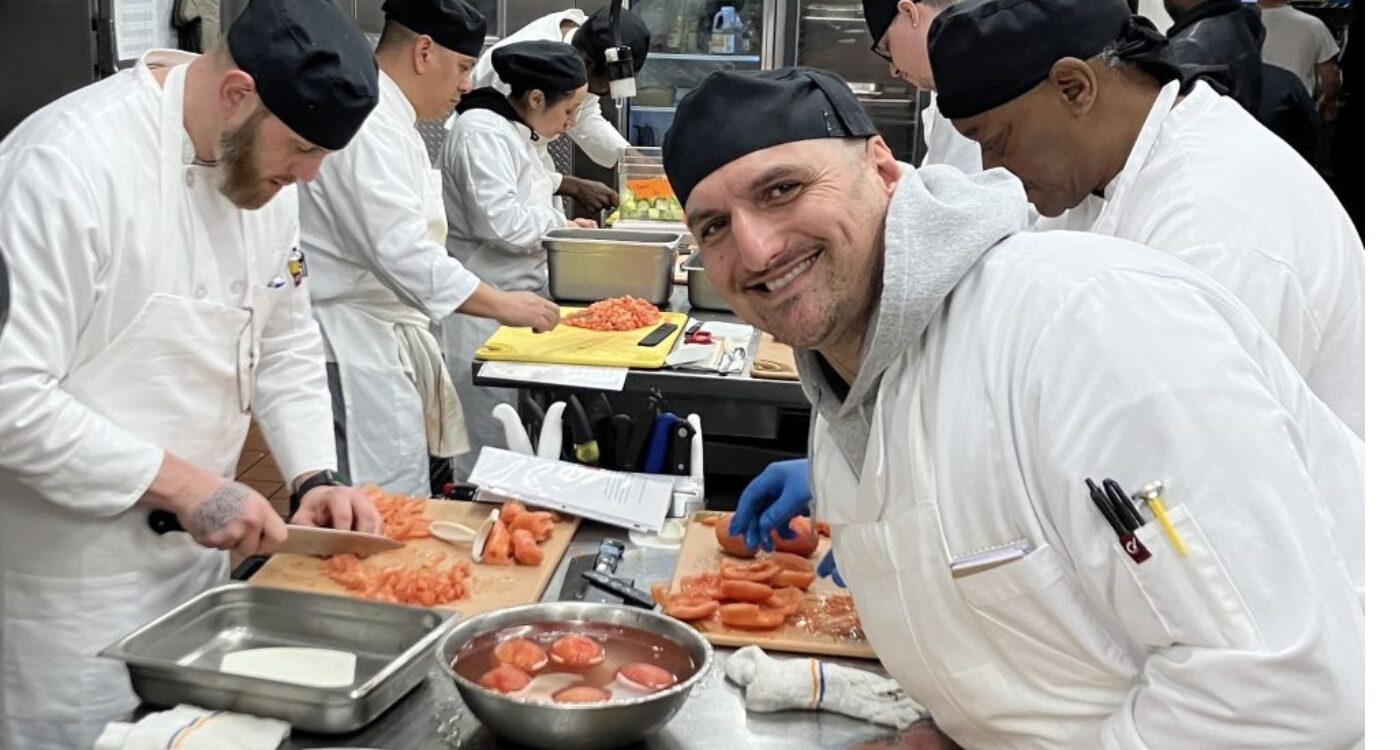 Man wearing chef coat and cap leans over kitchen counter smiling at camera