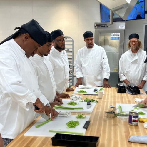 Culinary students in white coats gather at counter to dice celery