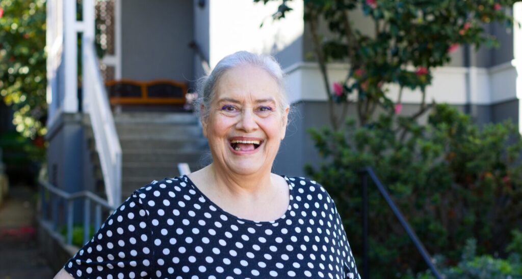 Older woman smiling in front yard with view of front porch steps and garden