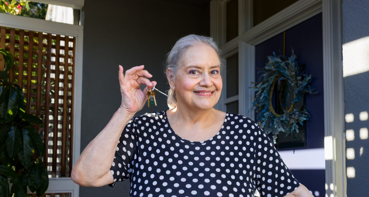 Older woman smiles while holding up key as she stands on front porch