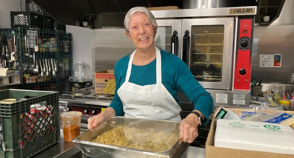 Woman with blue shirt and white apron holds warming tray of pasta