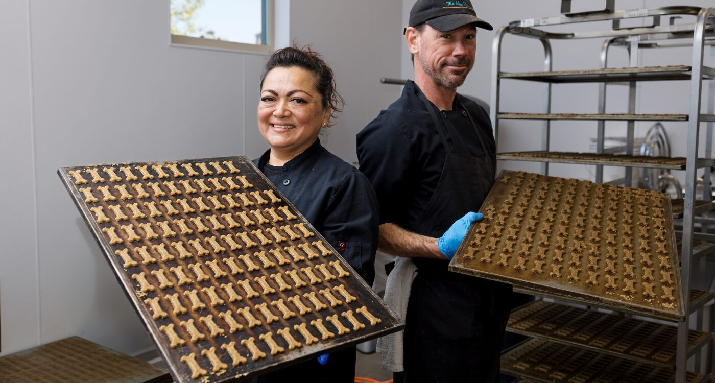Woman and man in black chef coats hold trays of dog treats ready for baking.