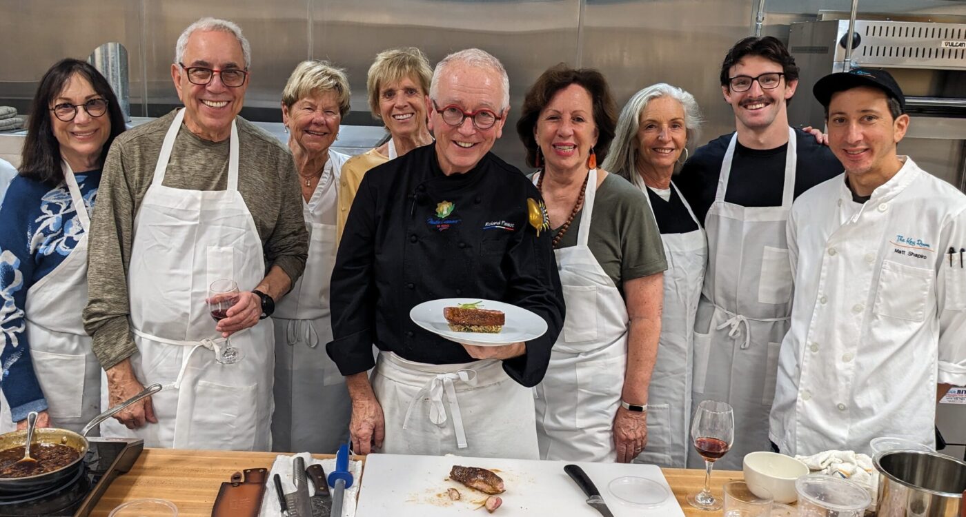 Chef in black coat stands with guests in white aprons at the prep counter