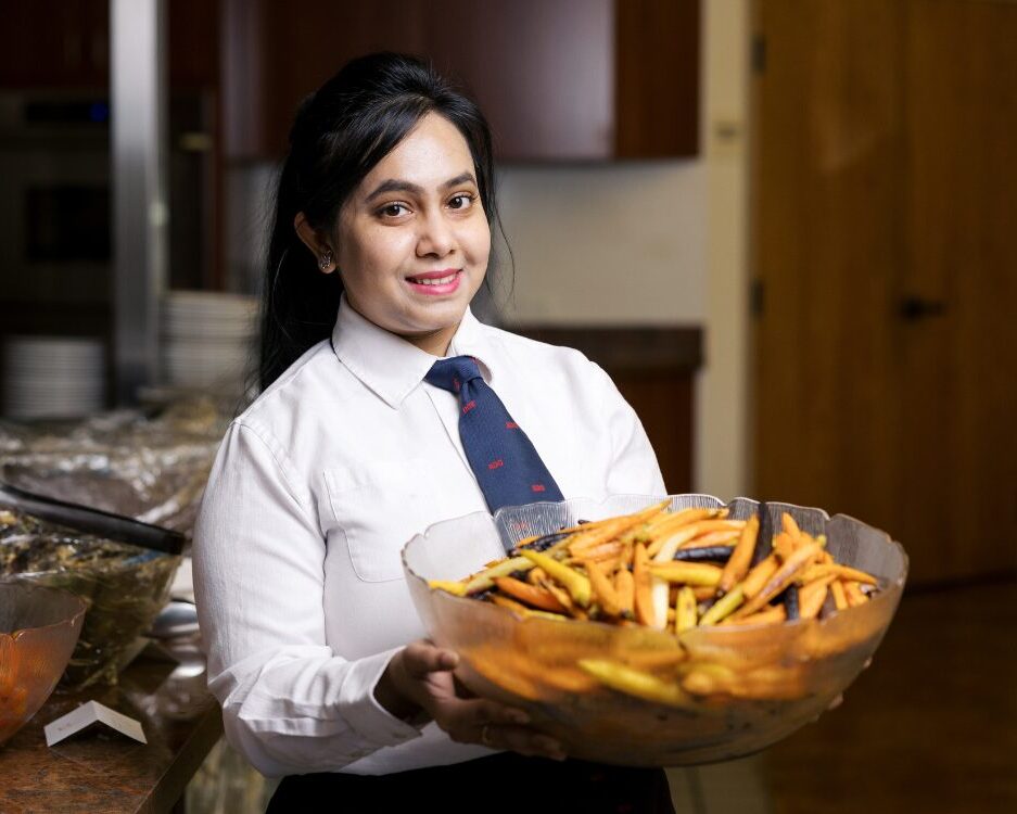 Woman wearing white shirt and blue tie holds bowl of roasted carrots