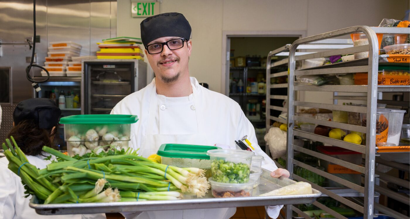 Man with glasses and white chef coat holds tray of scallions, chopped broccoli and boxes of mushrooms