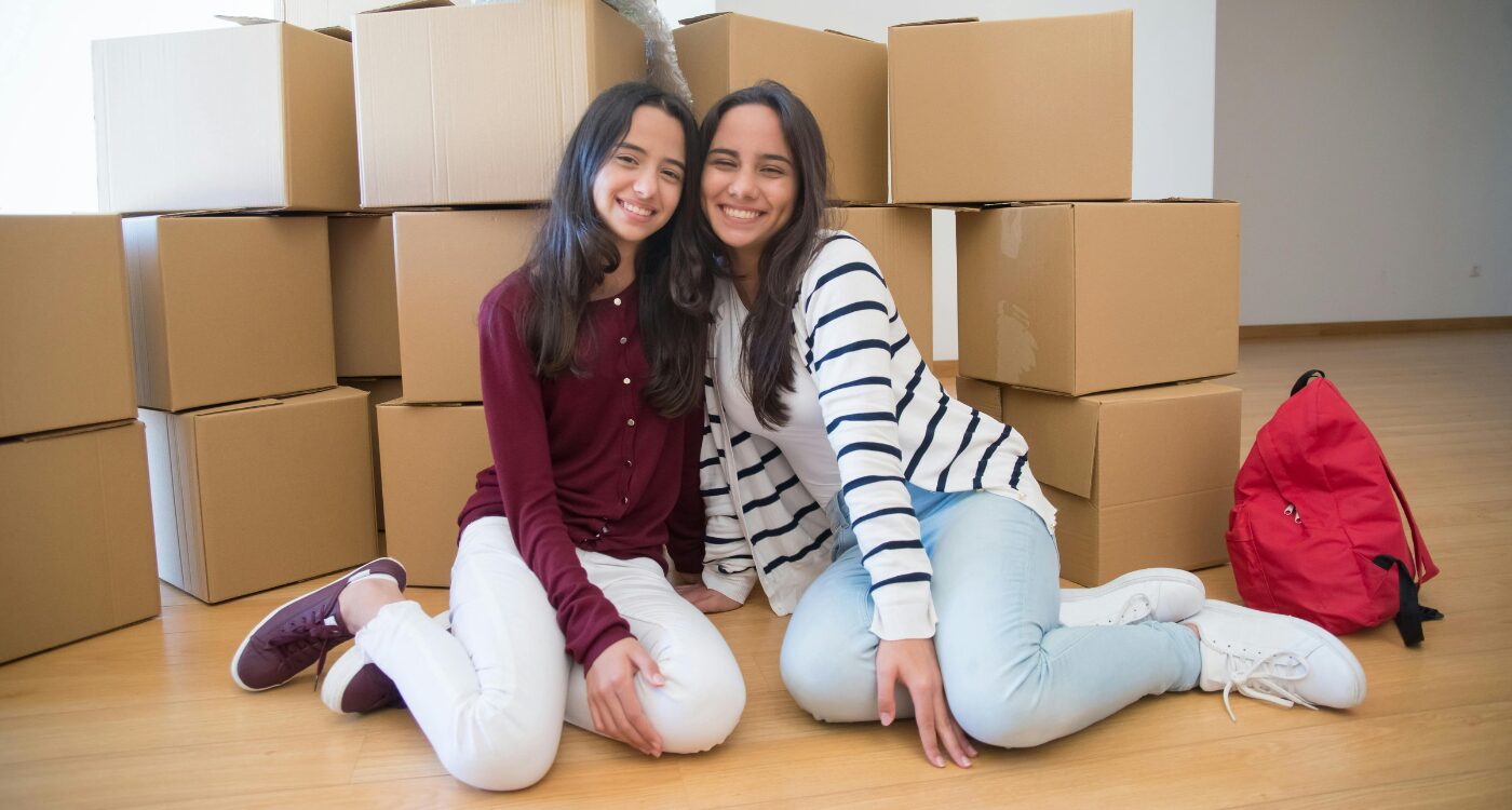 Two teen girls with long dark hair post in front of boxes with knapsacks