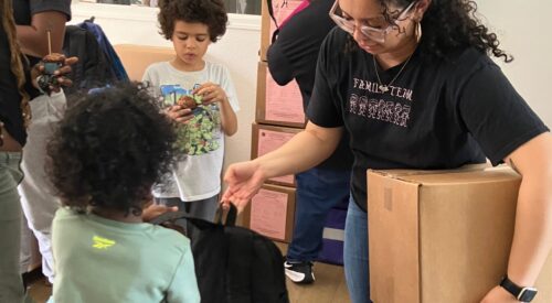 Woman in black T-shirt and glasses hands backpack to young boy.