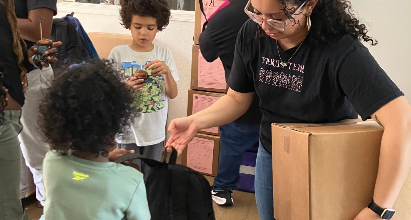 Woman in black T-shirt and glasses hands backpack to young boy.
